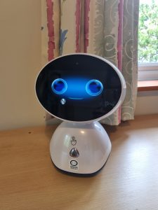 As small white desktop robot with an oval head black screen face and large blue round eyes sits on a wooden desk.