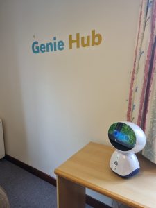 A small desktop robot with a cartoon style face and large eyes which blink sits on a wooden desk. A wall with the words Genie Hub sits behind the desk in a residential home at Anvil Court in Cornwall