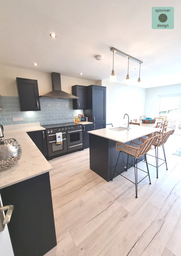 A modern open plan navy shaker kitchen with large central island in white marble, light grey wooden flooring and white walls is set off with bright modern artwork and a rustic oak dining table.